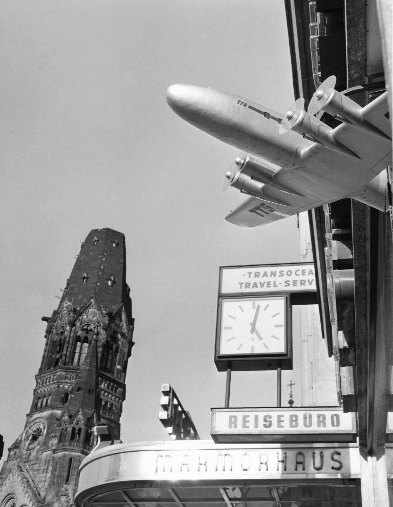 Das Schwarz-Weiß-Foto aus dem Jahr 1955 wurde von schräg unten aufgenommen. Im Vordergrund ragt, von rechts kommend, ein Flugzeugmodell ins Bild, das wohl an einer Häuserwand befestigt ist. Dahinter sind eine Uhr und mehrere Schilder mit den Aufschriften „Transocean Travel Service“, „Reisebüro“ und „Marmorhaus“ zu sehen. Links auf dem Bild ist, in etwas Entfernung, der Turm der Berliner Gedächtniskirche zu erkennen.