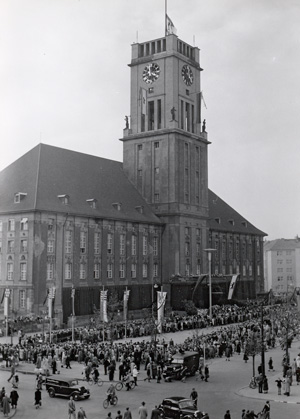 Die Schwarz-Weiß-Aufnahme aus dem Jahr 1951 zeigt das Rathaus Schöneberg in Berlin, das von 1949 bis 1991 Sitz des Regierenden Bürgermeisters war. Im Vordergrund ist der Rudolph-Wilde-Platz zu sehen, der 1963 nach John F. Kennedy benannt wurde. Einzelne Personen überqueren die Straße zum Platz, auch sind mehrere parkende Autos zu erkennen. Hinter dem Platz erhebt sich die mächtige Fassade des Rathauses mit seinem Glockenturm.